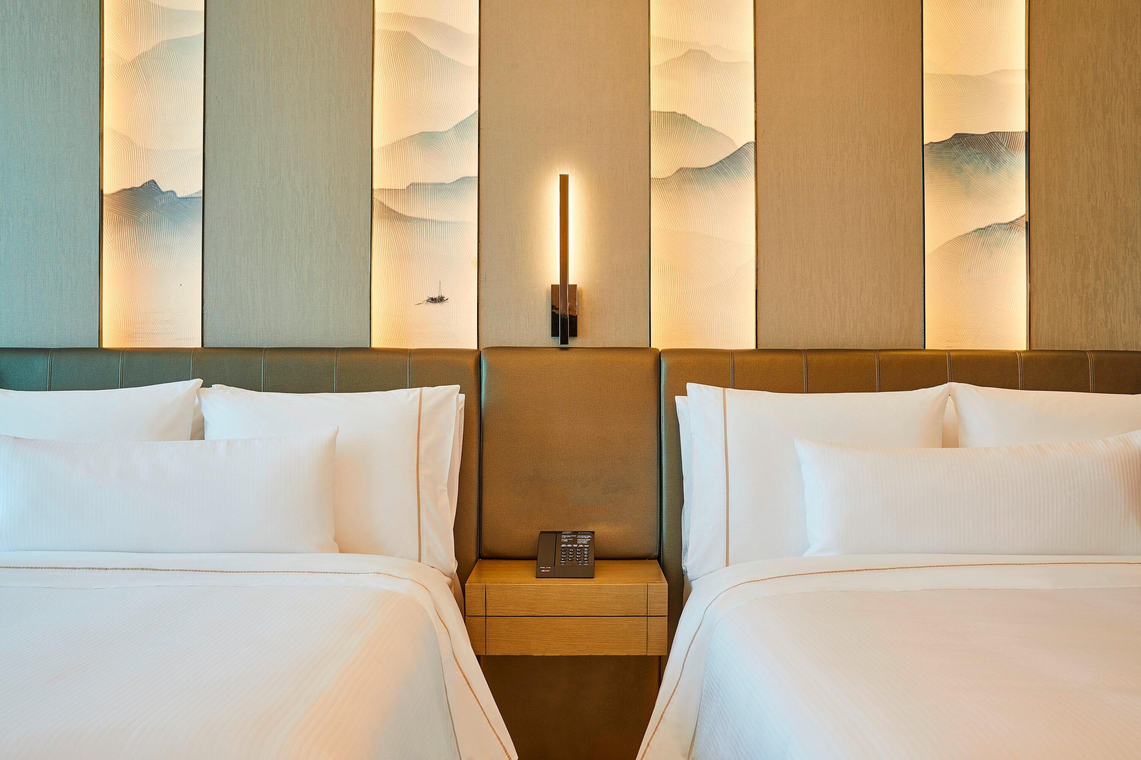 Quarto de hotel com duas camas, uma mesa de cabeceira com telefone e parede com design texturizado.
