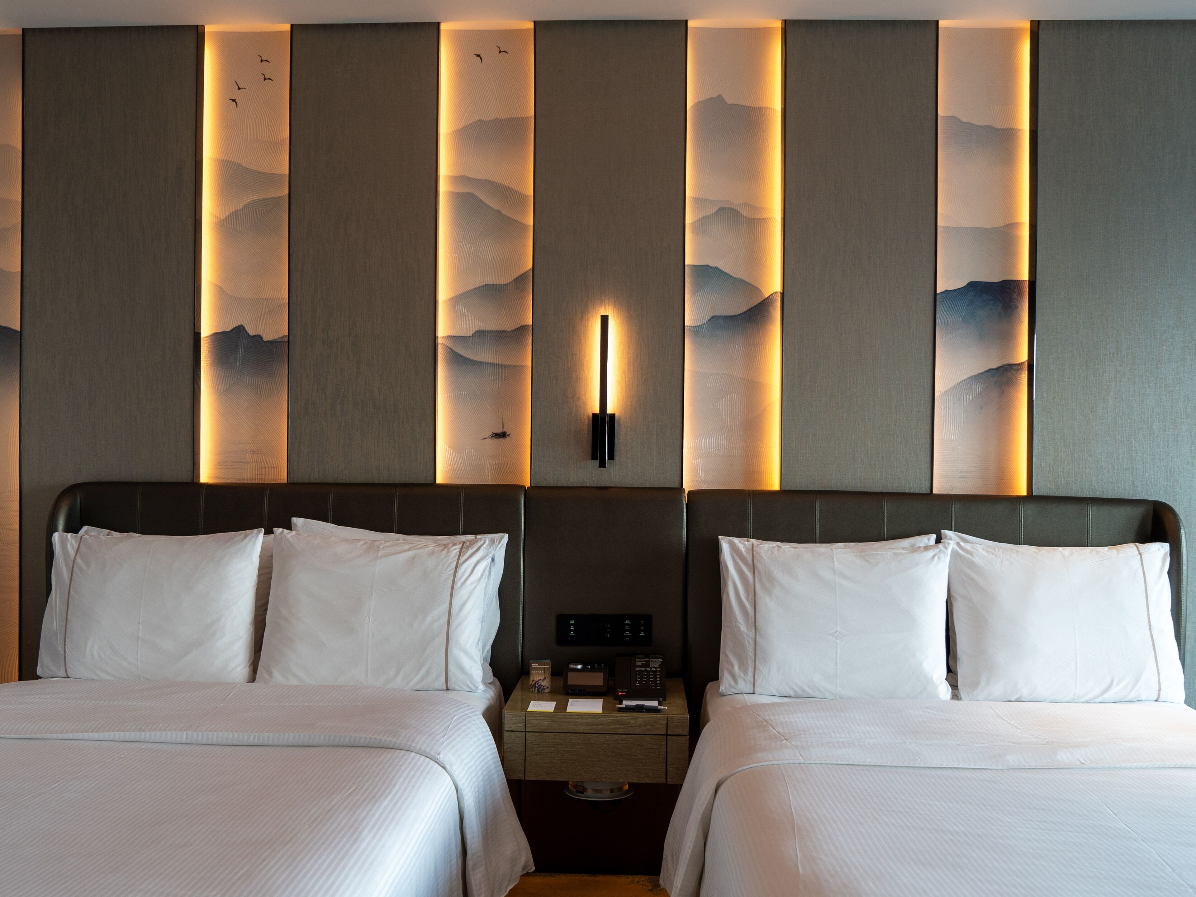 A hotel room with two beds, a nightstand with a phone and a lamp, and a wall with a textured design.