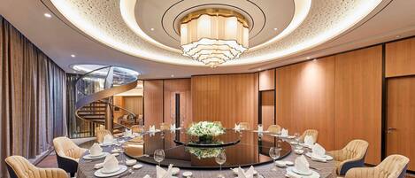 A round table with a central floral arrangement, surrounded by upholstered chairs, in a room with a modern chandelier and a spiral staircase.