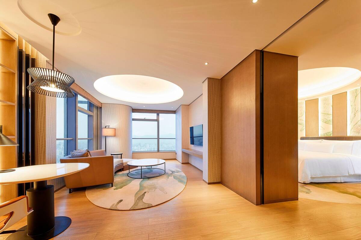 Ein modernes Hotelzimmer mit einem großen Bett, einer Sitzecke mit Sofa und rundem Tisch, einem großen Fenster mit Blick auf die Stadt und einer Decke mit runden Leuchten.