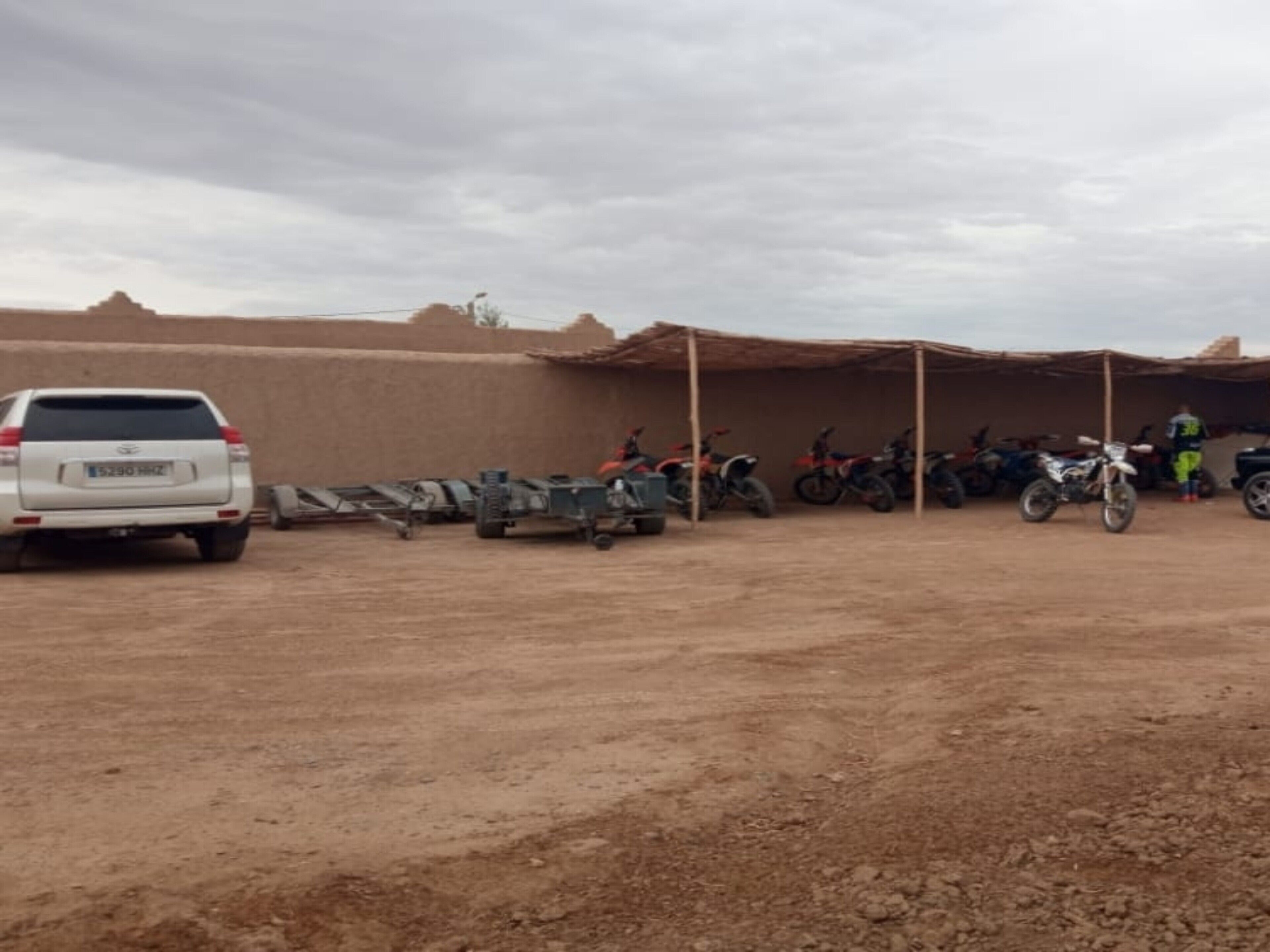 A dirt parking area with a white SUV and several motorcycles parked under a shelter.