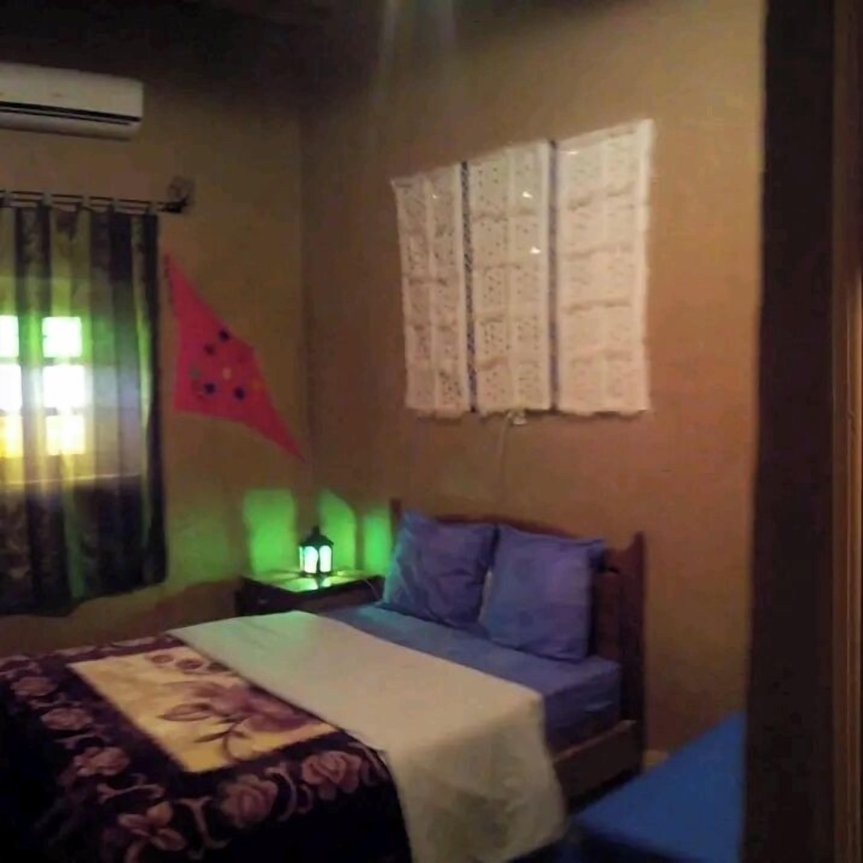 A bedroom with a bed, a nightstand, a wall-mounted lamp, and a window with curtains.