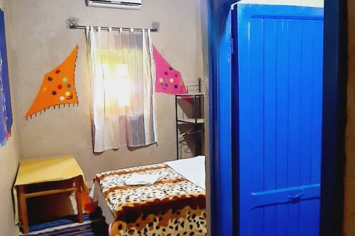 Una habitación con cama, una mesita, una silla, un ventanal con cortinas, un equipo de aire acondicionado y una puerta azul.