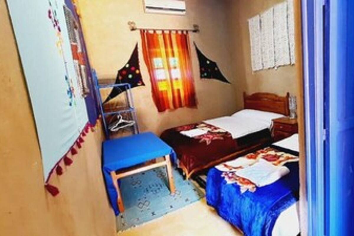 Habitación pequeña y funcional con cama, escritorio y silla. Cuenta con ventana con cortinas, aire acondicionado instalado en la pared y puerta que da a otra habitación.