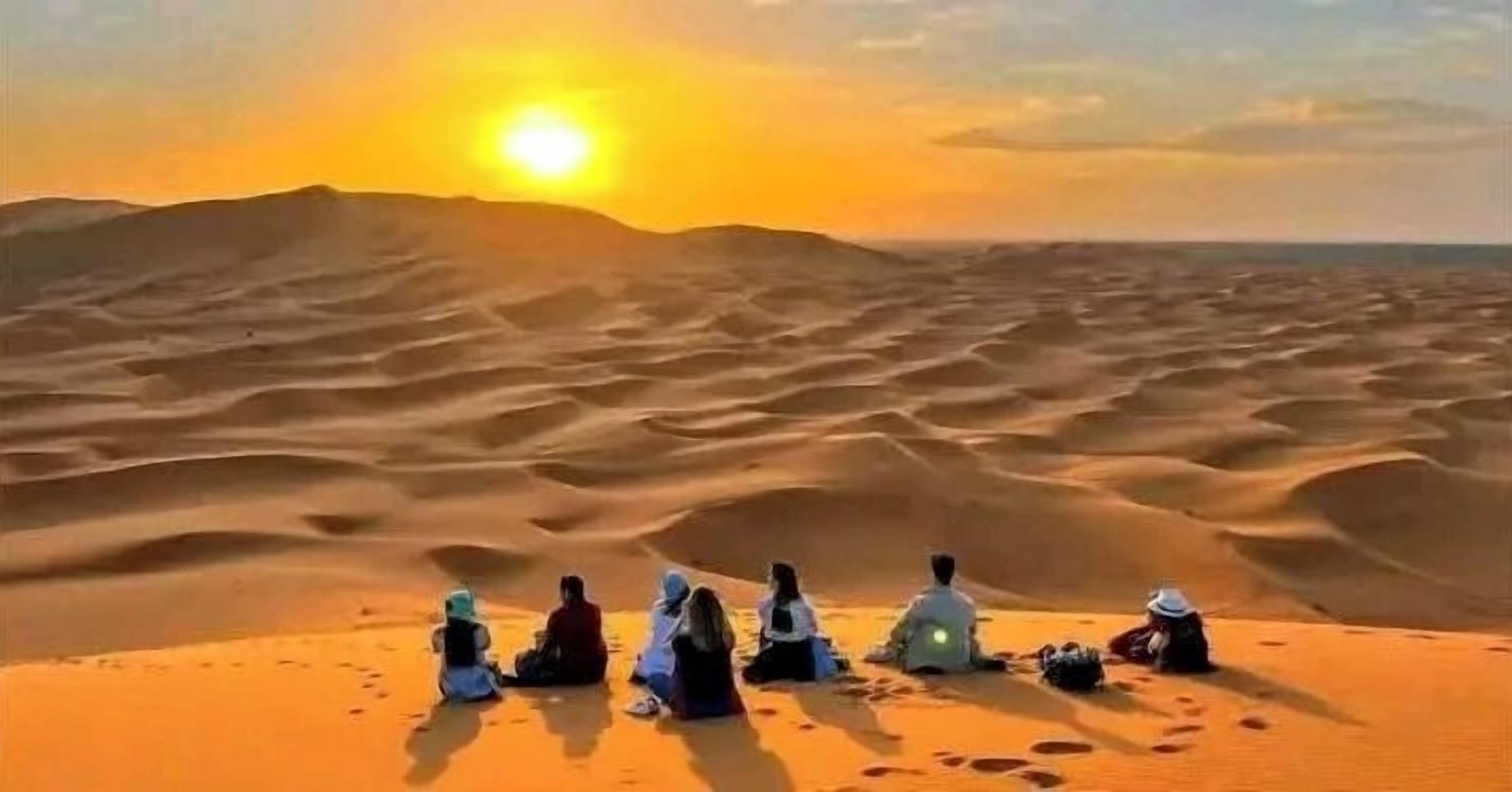 A group of people sitting on a sand dune at sunset.