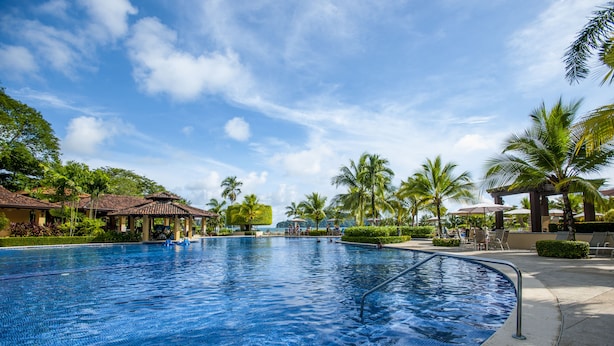 A large outdoor swimming pool with clear blue water, surrounded by palm trees and lounge chairs. - Los Suenos Resort Miramar 5D (Jacó)