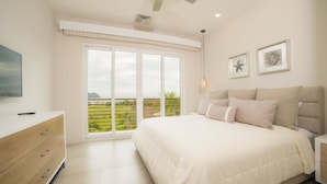 A bedroom with a large bed, a dresser, a TV, and a balcony with a view of greenery. - Los Suenos Resort Miramar 5D (Jacó)
