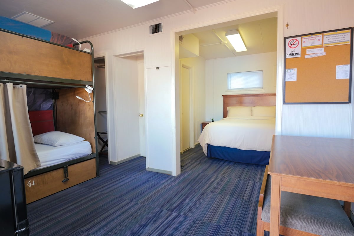 Una habitación de hotel con literas, una cama individual, una mesa de madera y un tablón de anuncios.