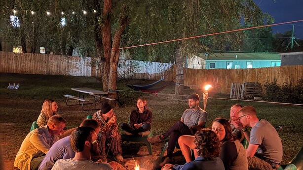 A group of people seated around a campfire in an outdoor setting with a wooden fence and trees in the background. - RAD Hostel (Colorado Springs)