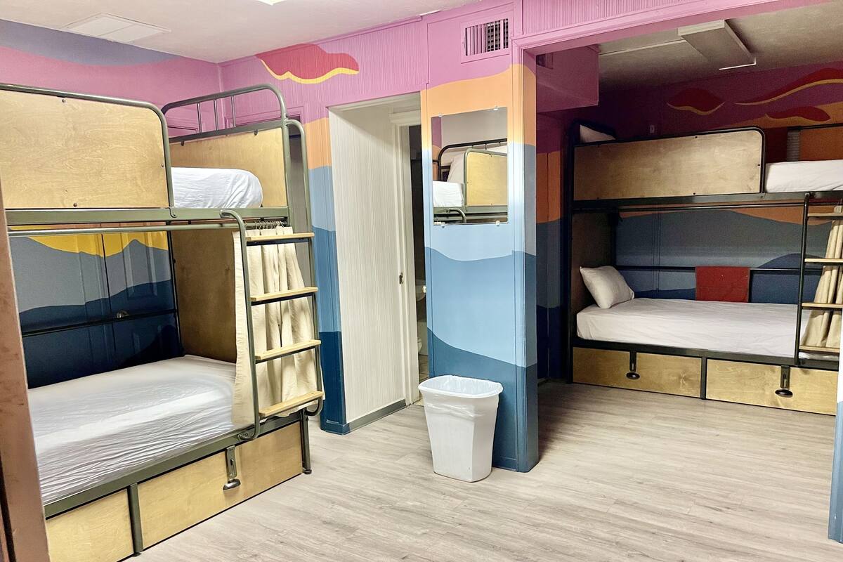 Un dormitorio con literas, una escalera, un cubo de basura y una puerta que da a otra habitación.