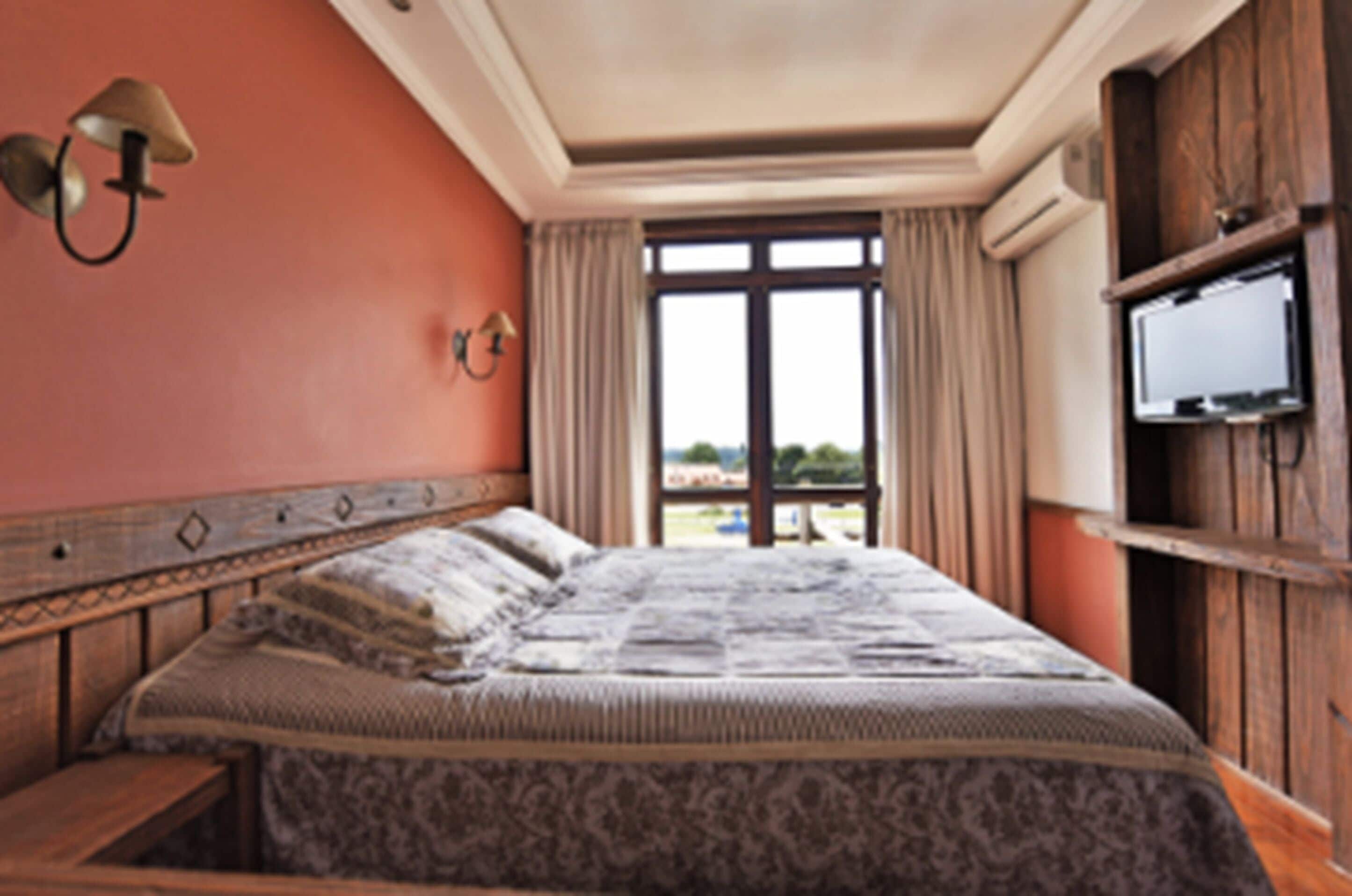 A bedroom with a large bed, a wooden headboard, a wall-mounted TV, and a window with curtains.