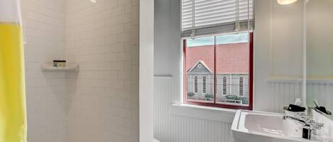 Un baño con paredes de azulejos blancos, una ventana con vista a un edificio rojo, un lavamanos blanco y una zona de ducha.