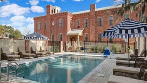 A pool area with lounge chairs and umbrellas, a brick building with arched windows, and a clear blue sky. - Amelia Schoolhouse Inn (Fernandina Beach)
