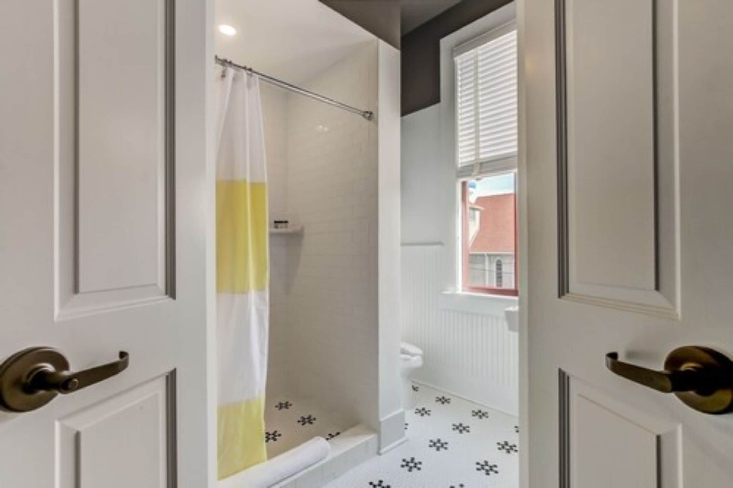 Un baño con ducha, paredes de azulejos blancos, cortina de ducha amarilla y blanca, y una ventana con persianas.