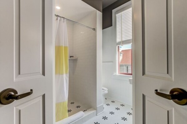A bathroom with a shower, white tiled walls, a yellow and white shower curtain, and a window with blinds.
