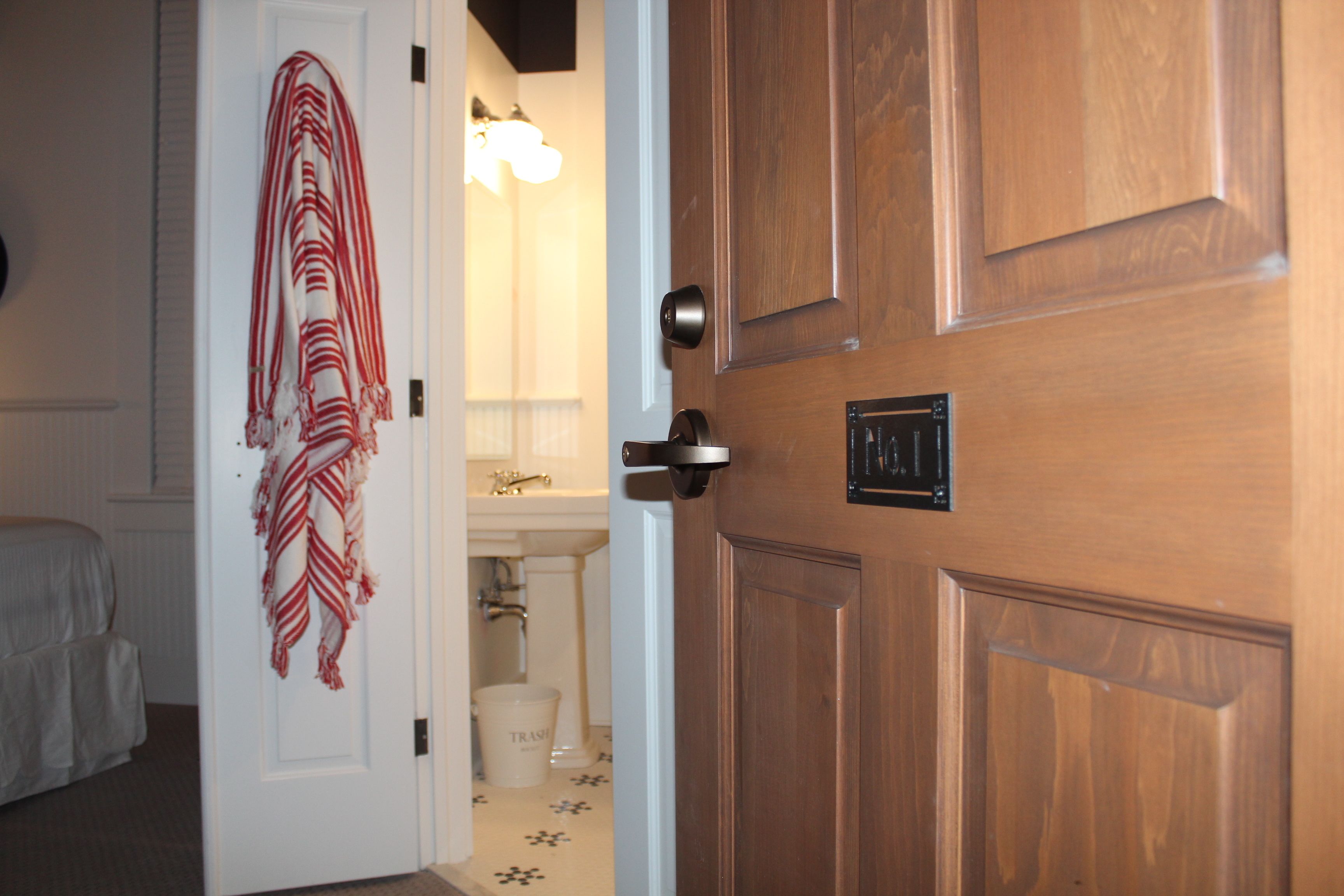 A hotel room with a wooden door, a white door with a red and white striped towel hanging on it, and a bathroom visible through the door.