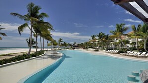 Ein klares, blaues Schwimmbecken mit gewölbter Kante, umgeben von Palmen und Liegestühlen, mit Strand und Hotelgebäuden im Hintergrund.