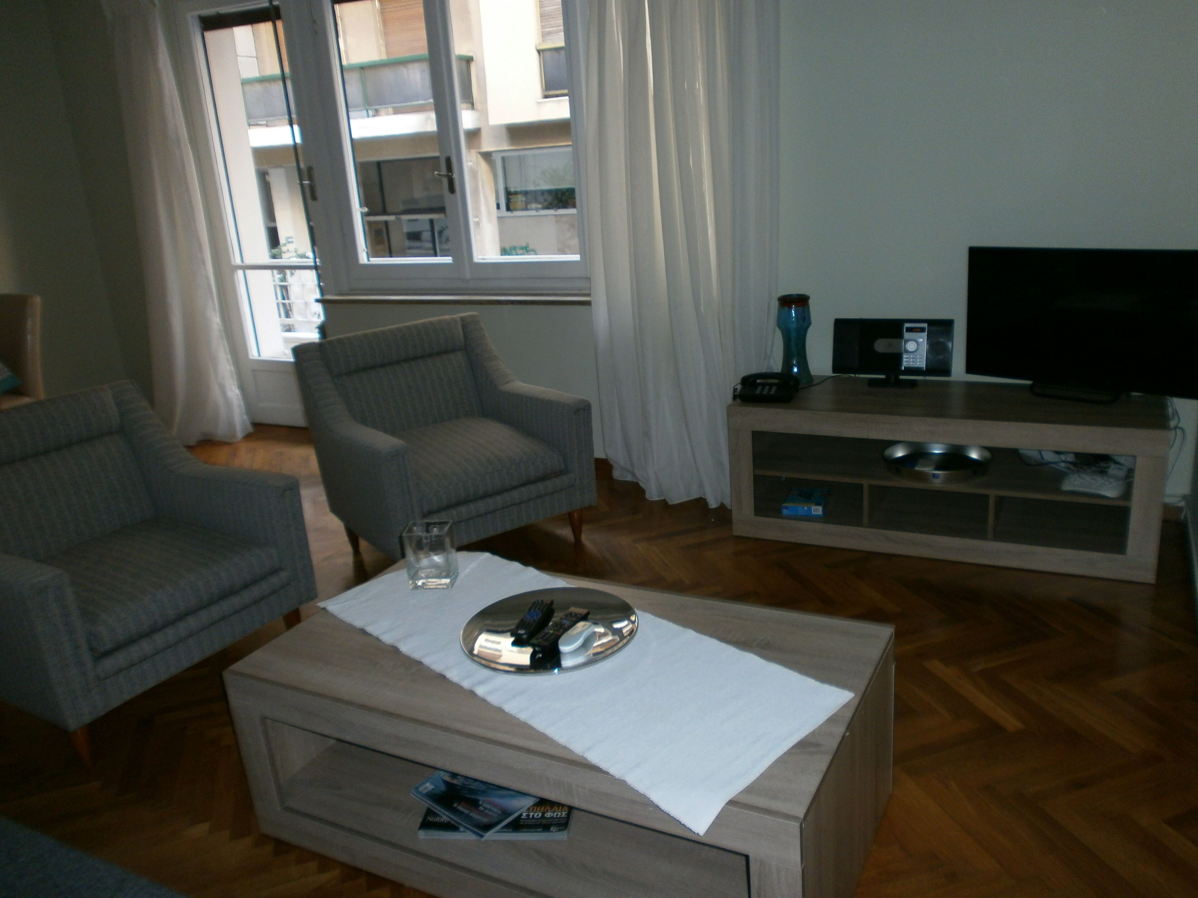 A living room with a sofa, armchair, coffee table, TV stand, and a view of an apartment building through the window.