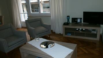 A living room with a sofa, armchair, coffee table, TV stand, and a view of an apartment building through the window.