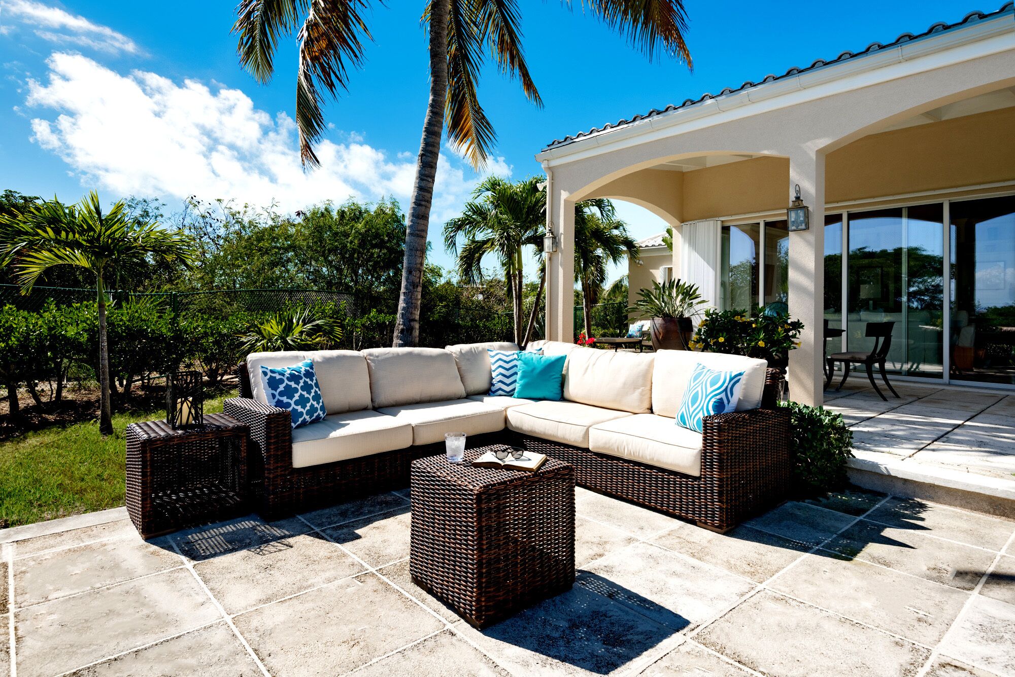 A covered outdoor seating area with a beige sectional sofa, wicker side tables, and a glass door leading to a patio.