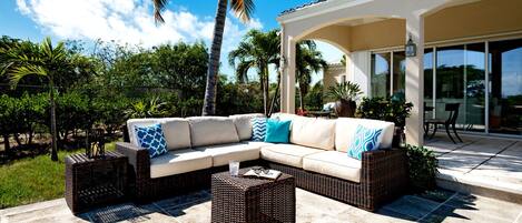 A covered outdoor seating area with a beige sectional sofa, wicker side tables, and a glass door leading to a patio.