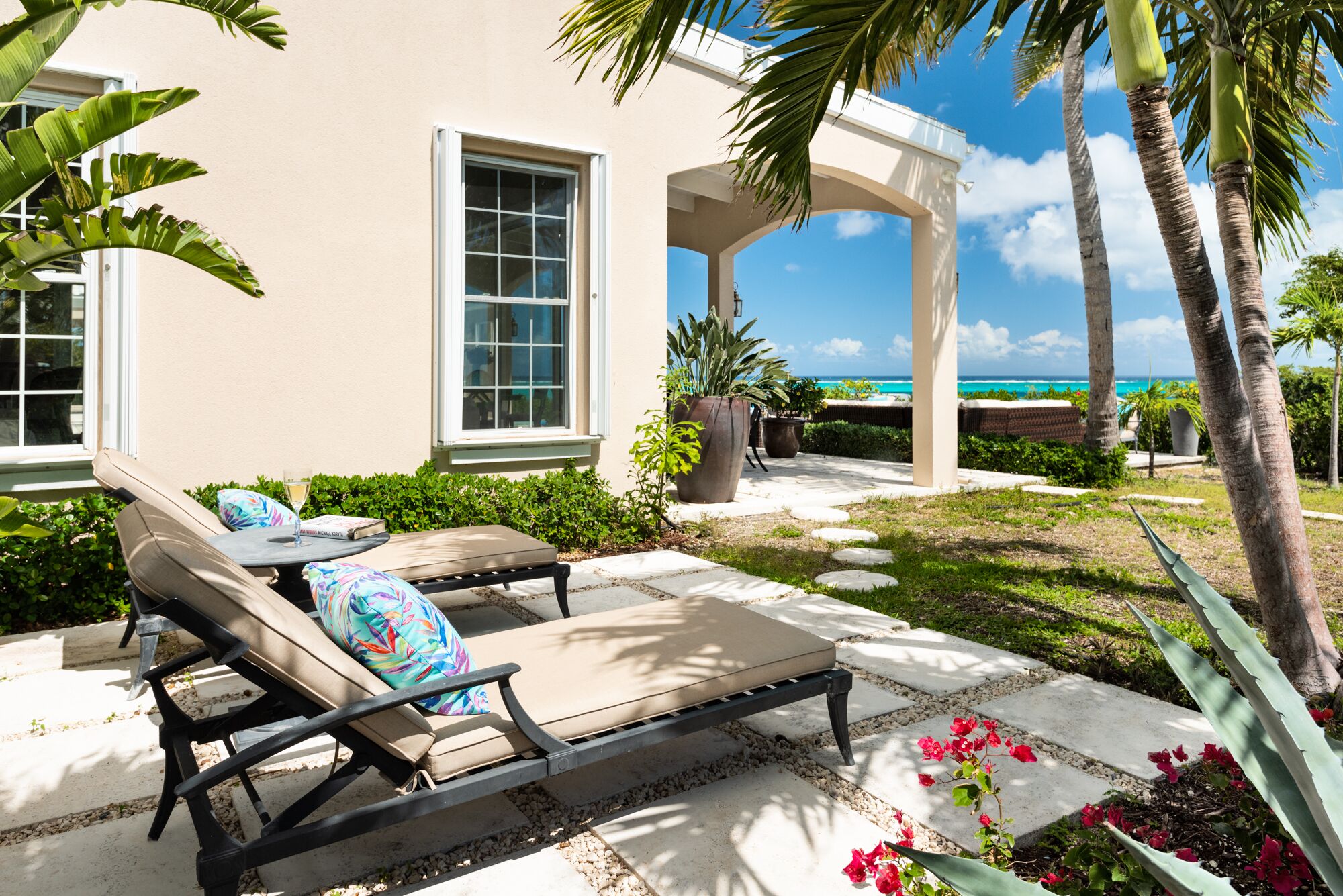 A sunlit patio with lounge chairs, a large potted plant, and a view of the ocean.