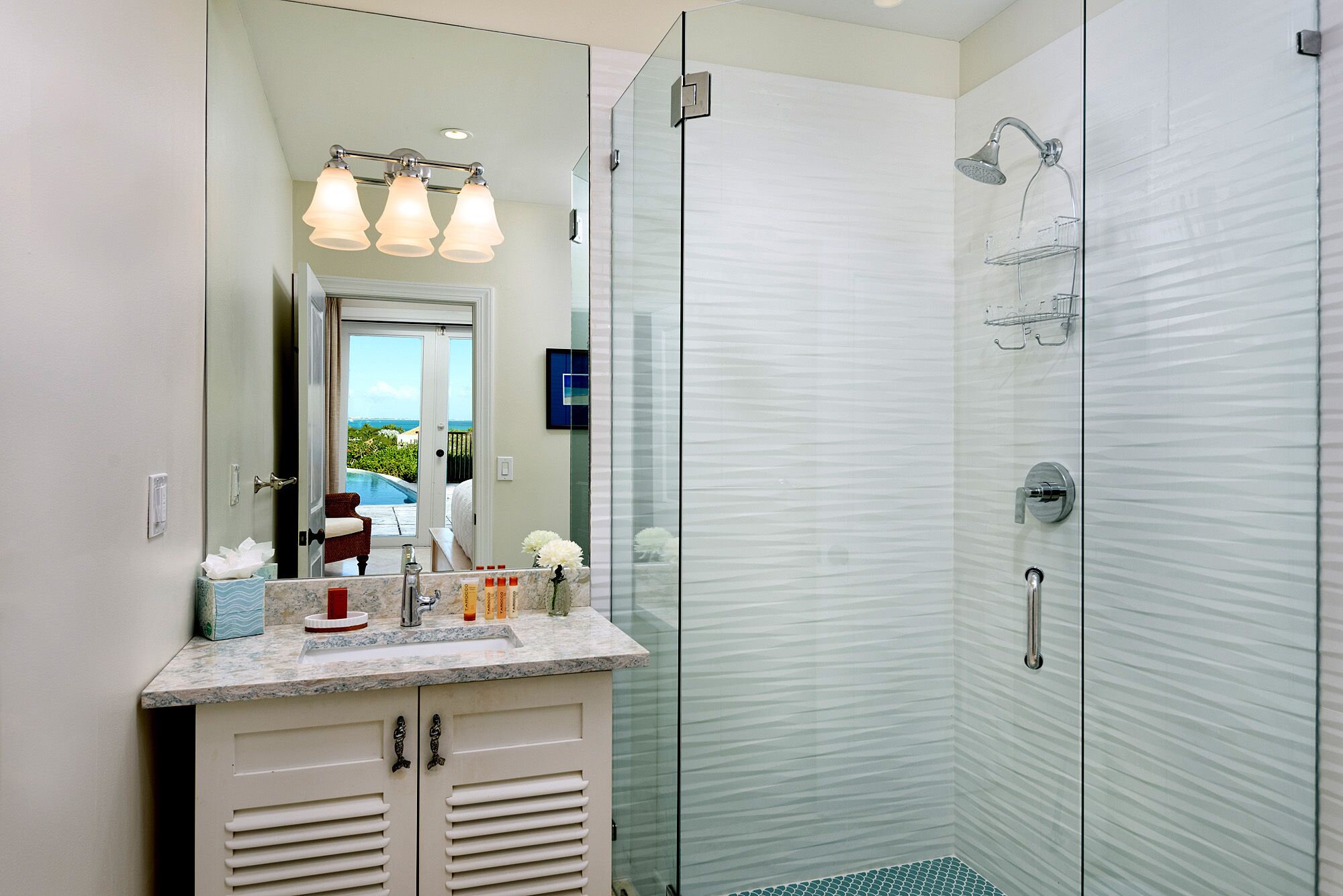 A modern bathroom with a glass shower enclosure, a vanity with a marble countertop, and a view of a pool and greenery through the glass door.