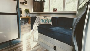 A compact living space with a kitchenette, a sofa with a pillow labeled "FLORIDA," and a window with curtains. - With Oasis Shared Backyard (St. Petersburg)