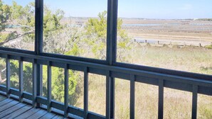A view from a covered porch with a railing, overlooking a coastal area with a bridge and open fields.