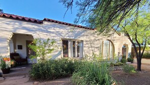 Una casa con techo de tejas, un porche con una silla y un jardín con diversas plantas.