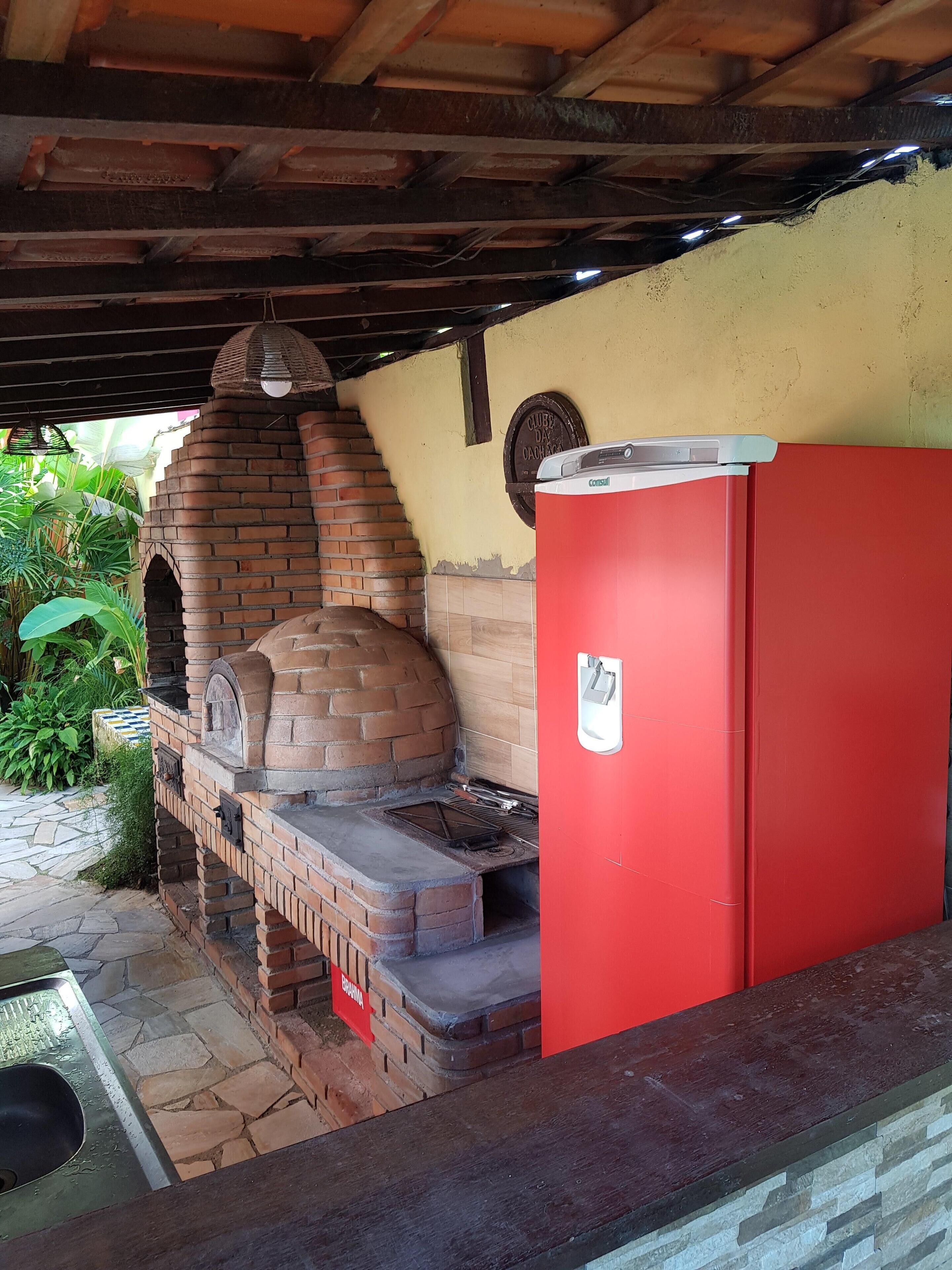 Un horno de ladrillo bajo una zona cubierta, con un refrigerador rojo cercano.