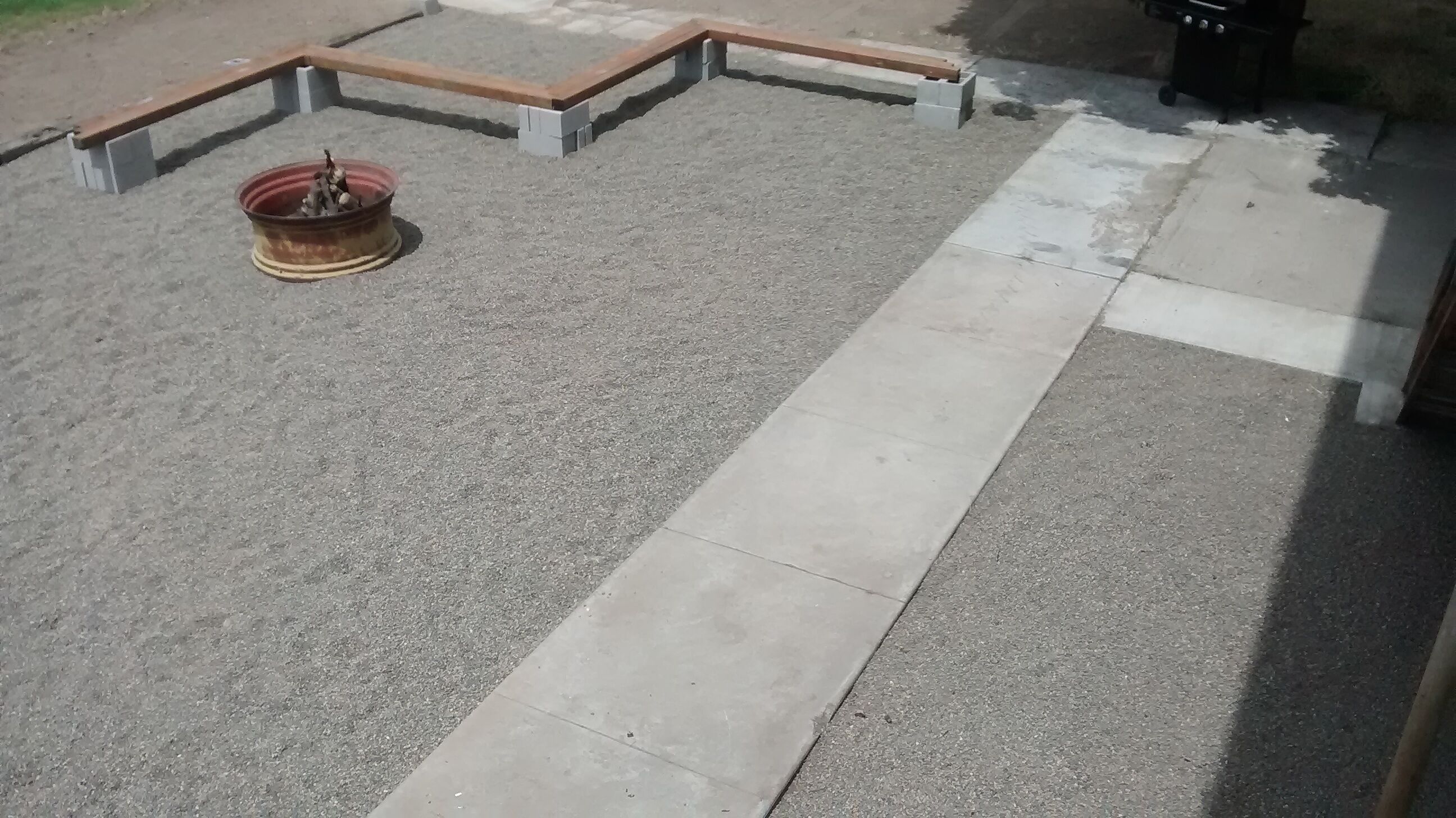 Un chemin en béton mène à un espace agrémenté de bancs en bois et doté d’un foyer.