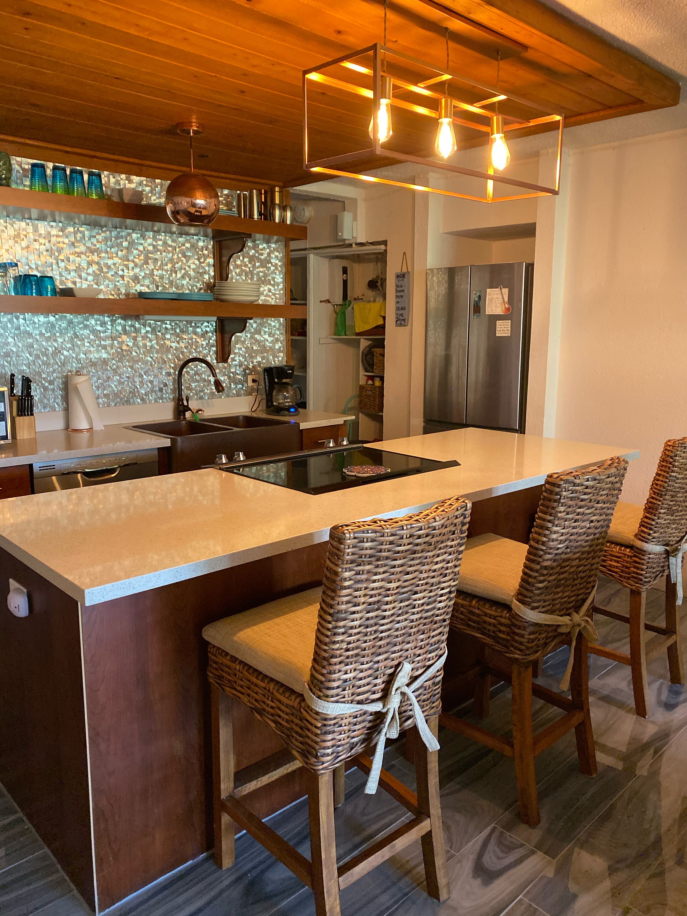 Een moderne keuken met een centraal eiland, barstoelen, een roestvrijstalen koelkast en een hangende lamp.