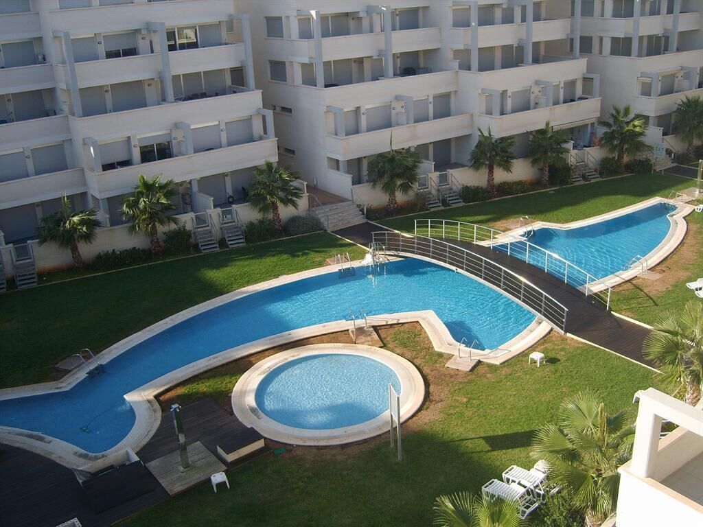 A multi-story hotel with a pool area, including a large rectangular pool and a smaller circular pool, surrounded by palm trees and a well-maintained lawn.