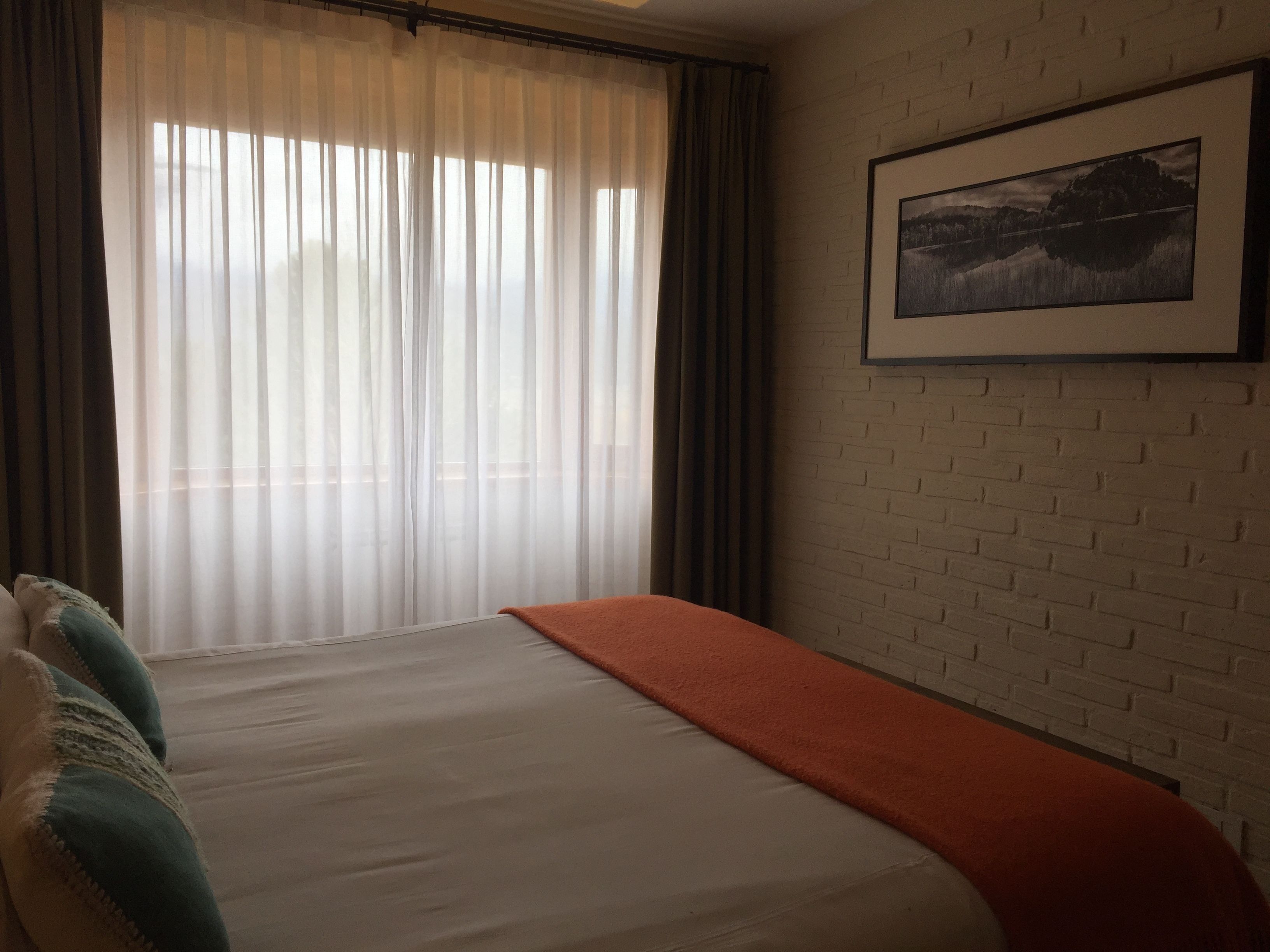 A hotel room with a bed, a window with sheer curtains, a brick wall, and a framed picture on the wall.