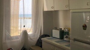 Eine kleine Küche mit weißen Schränken, einem Kühlschrank, einem Fenster mit transparenten Vorhängen und einer Flasche auf der Fensterbank.