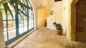 A corridor with stone flooring, a potted plant, and a view through a window with blue shutters. - Charming, 300 year old traditional Farmhouse with private pool (Zebbug)