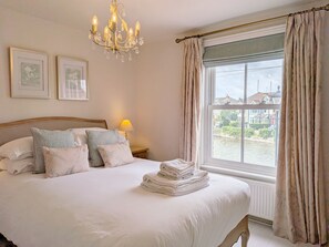 A neatly made bed with pillows, a chandelier, a lamp, and a view of a canal through a window. - Compass Cottage , Emsworth  -  a cottage that sleeps 4 guests  in 2 bedrooms (Emsworth)
