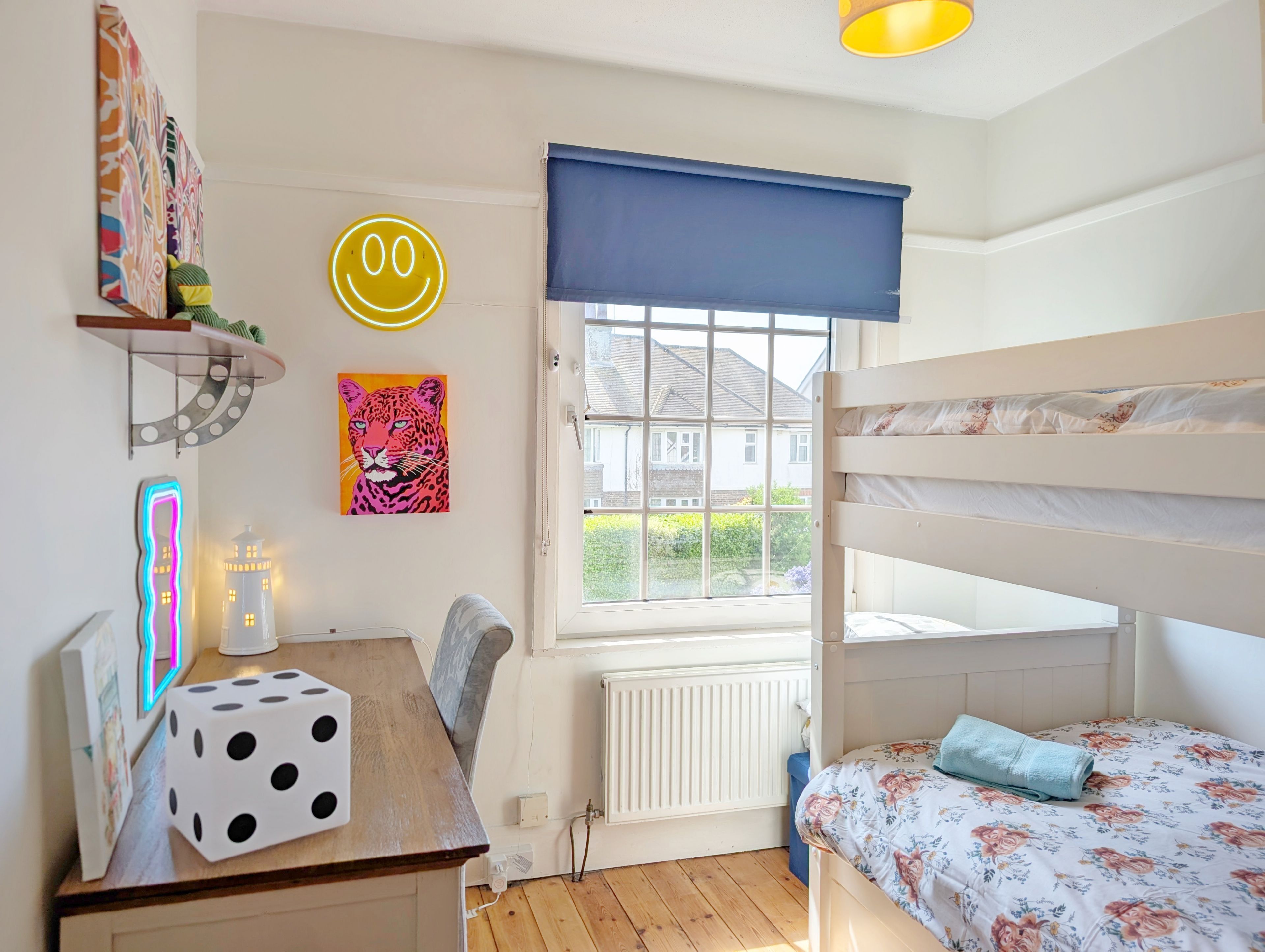A child's bedroom with bunk beds, a desk with a dice, a radiator, and a window with a view of houses.