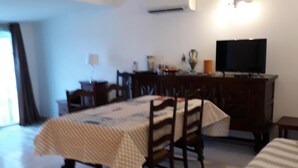 Zona de menjador amb taula coberta per un mantell a cuadros, cadires de fusta i un televisor sobre un suport.