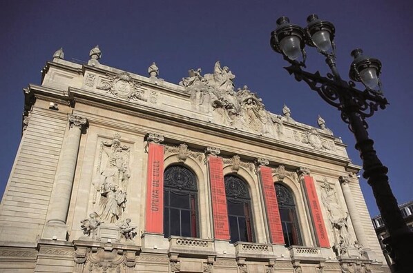 Fachada clásica de un edificio con esculturas ornamentales, una estatua destacada y banderines rojos.