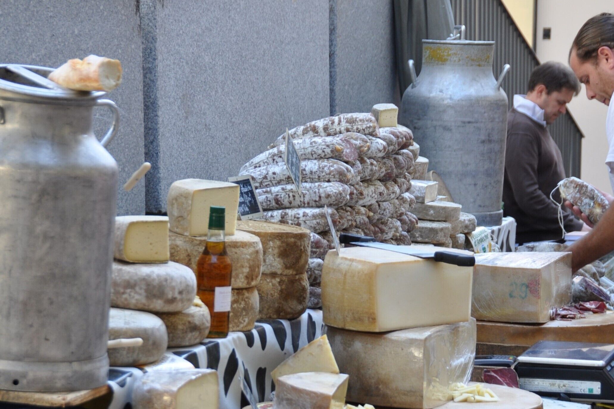 A cheese market stall with various cheeses, a large metal container, and a weighing scale.