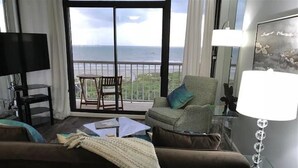 Una sala de estar con sofá, una mesa de centro, televisión y vista al océano a través de grandes ventanales.