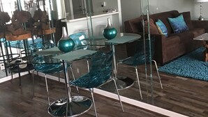 Una sala de estar moderna con una mesa de comedor de vidrio, sillas color turquesa y un sofá marrón con cojines azules.