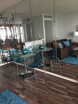 A modern living room with a glass dining table, teal chairs, and a brown sofa with blue cushions.