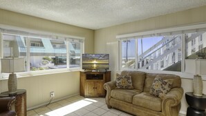 A living room with a beige couch, a television on a wooden stand, and large windows offering a view of a building and a beach.