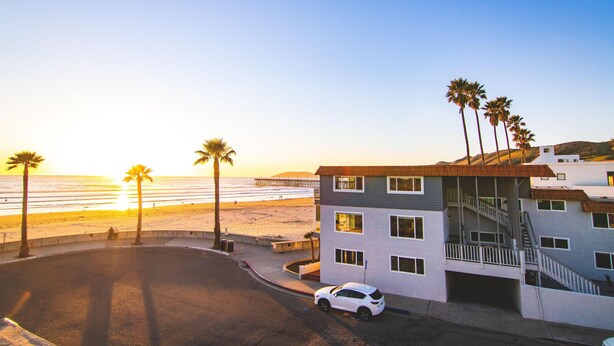 A modern multi-story building with a parking area, adjacent to a beach with palm trees and a view of the ocean.