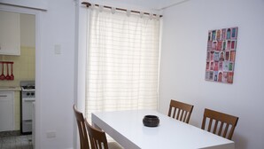 A dining area with a white table and wooden chairs, a window with a white curtain, and a kitchen visible in the background. - Comfortable Apartment in Palermo (Buenos Aires)