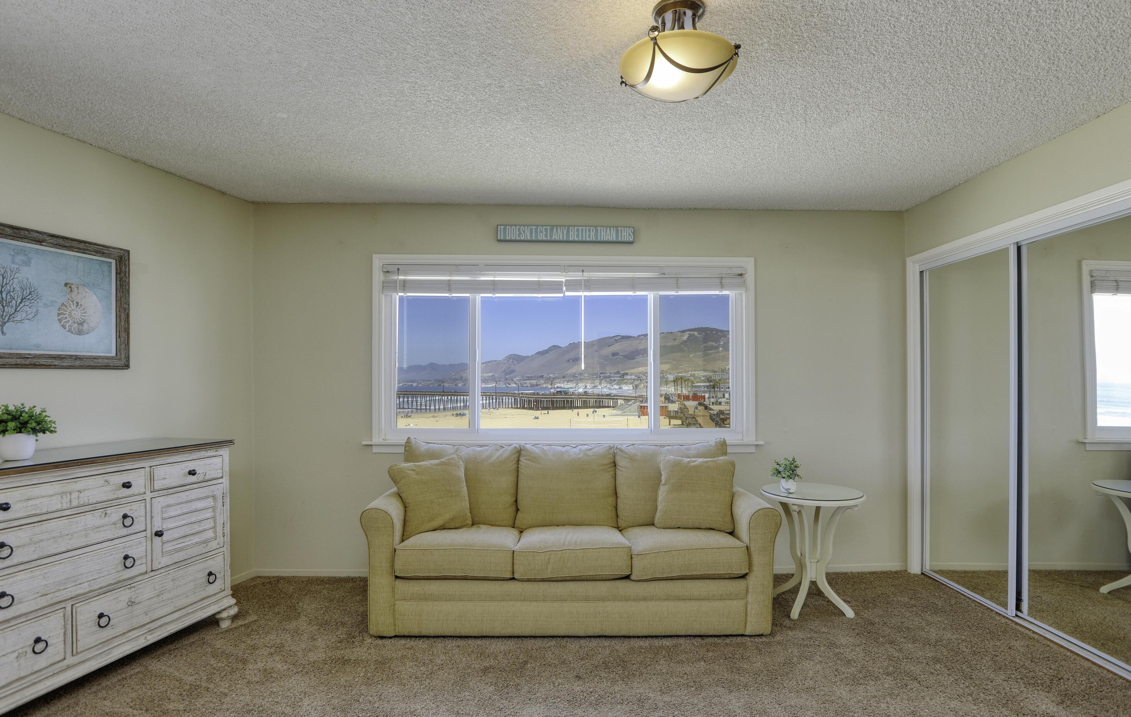 A room with a beige sofa, a small round table, a dresser, a large window with a view of mountains, and a ceiling light fixture.