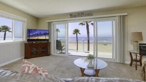 Una sala de estar con un televisor grande, una mesa de vidrio, un sofá y vistas a la playa a través de puertas correderas amplias.